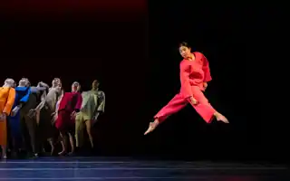 En samling dansare i färgglada kläder, i förgrunden hoppar en dansare i rött.