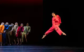 A group of dancers in colorful clothes, in the foreground a dancer in red jumps.