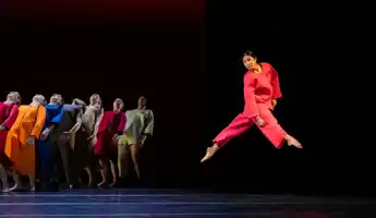 En samling dansare i färgglada kläder, i förgrunden hoppar en dansare i rött.