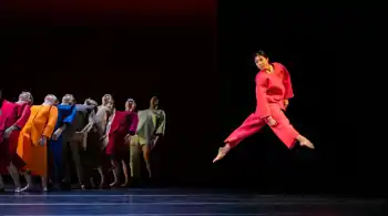 En samling dansare i färgglada kläder, i förgrunden hoppar en dansare i rött.