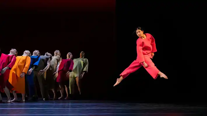 En samling dansare i färgglada kläder, i förgrunden hoppar en dansare i rött.