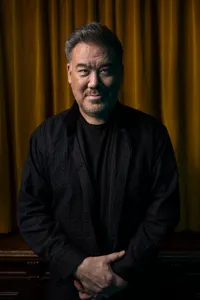 En mörkklädd man står med korslagda händer framför ett sammetsdraperi.