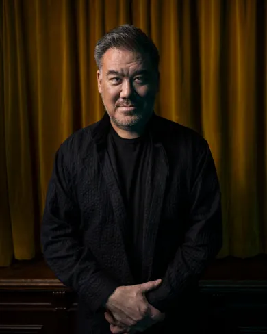 A man dressed in dark clothes stands with his hands folded in front of a velvet curtain.