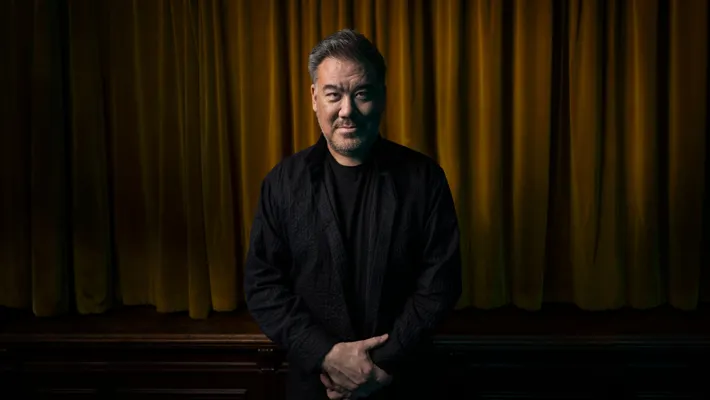 A man dressed in dark clothes stands with his hands folded in front of a velvet curtain.