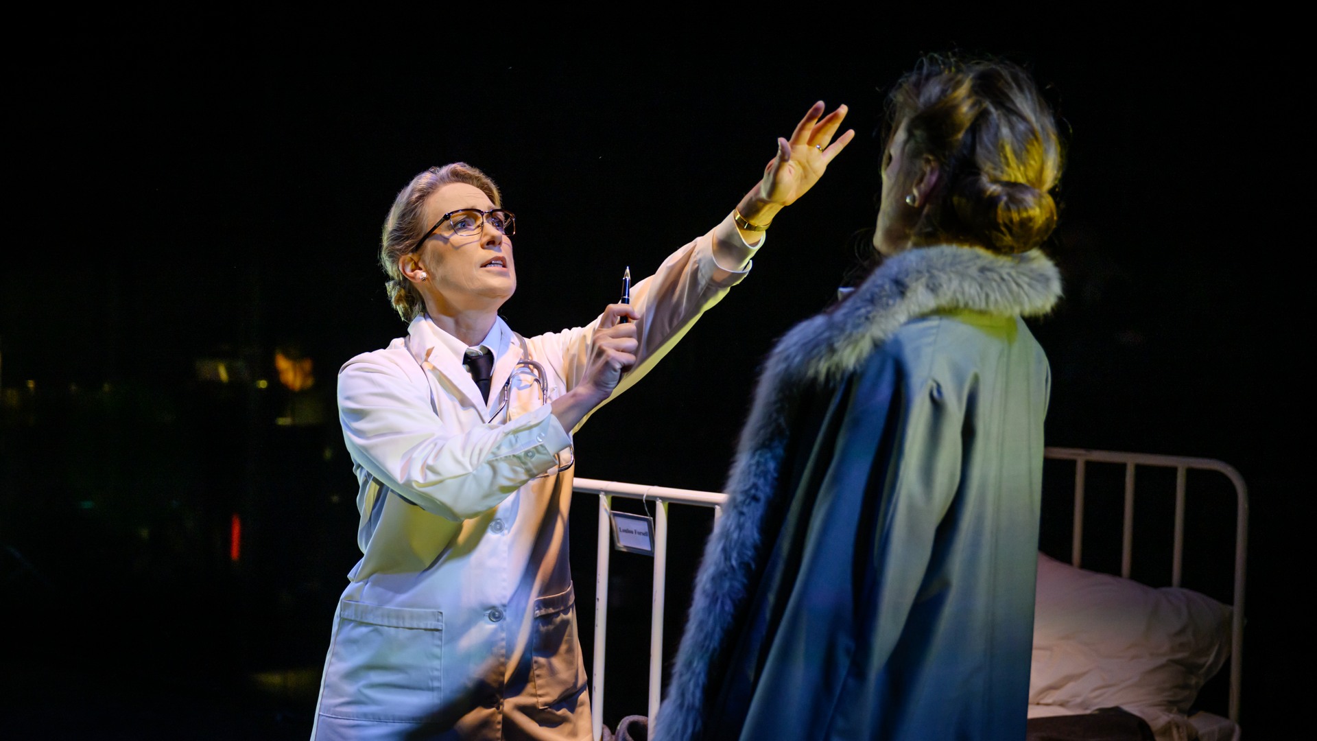 A woman in a doctor's coat extends her arm towards a woman in an overcoat, with a hospital bed in the background.