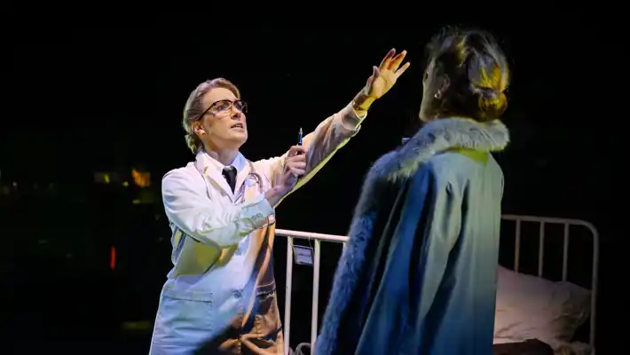 En kvinna i läkarrock sträcker ut sin arm mot en kvinna i överrock, i bakgrunden en sjukhussäng.