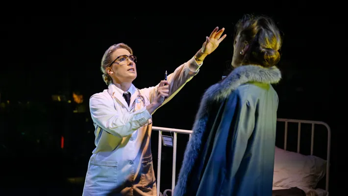 A woman in a doctor's coat extends her arm towards a woman in an overcoat, with a hospital bed in the background.