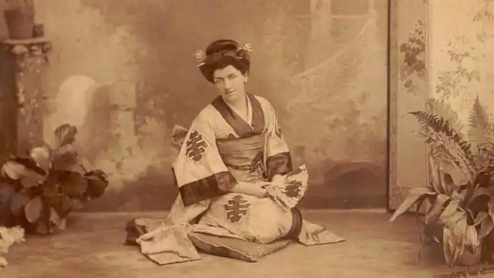 En kvinna sitter, iklädd en kimono, i handen en solfjäder.