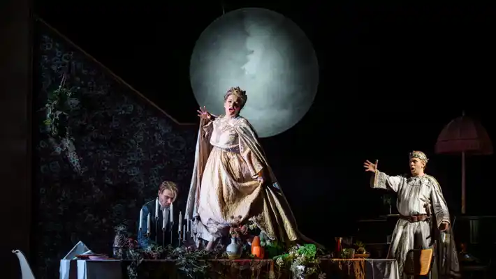 A woman stands on a set table wearing a crown, behind her a large moon and two men.