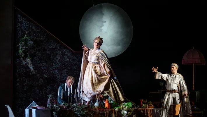 En kvinna står på ett dukat bord iförd en krona, bakom henne en stor måne och två män.