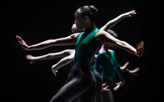 Dansare med gröna kläder mot en mörk bakgrund, de gör rörelser med sina armar.