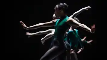 Dansare med gröna kläder mot en mörk bakgrund, de gör rörelser med sina armar.