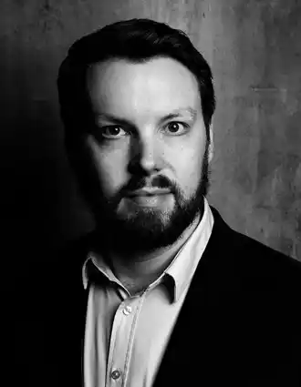 A close-up of a man with dark hair and a beard wearing a light shirt and a dark jacket.