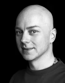 Close-up photo in black and white of a man wearing a dark sweater and a necklace.