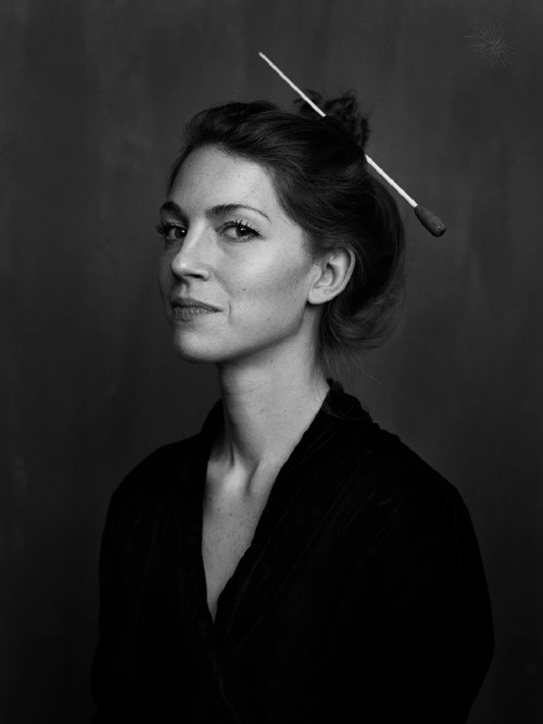 Portrait image of a woman in half profile, she is wearing a dark jacket and her hair is tied up with a conductor's baton.