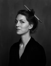 Portrait image of a woman in half profile, she is wearing a dark jacket and her hair is tied up with a conductor's baton.