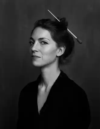 Portrait image of a woman in half profile, she is wearing a dark jacket and her hair is tied up with a conductor's baton.