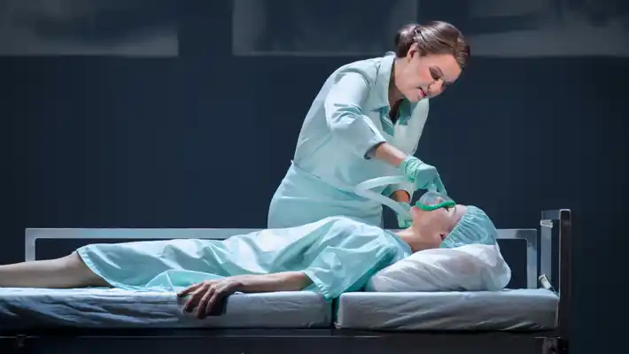 En kvinna i sjukhuskläder ligger på en bår, en sjuksköterska ger henne syrgas med en mask.