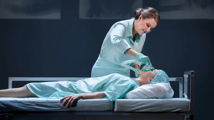 A woman in hospital clothes lies on a stretcher, a nurse gives her oxygen with a mask.