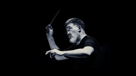 A man in a black short-sleeved shirt with glasses and a conductor's baton in his hand against a dark background.