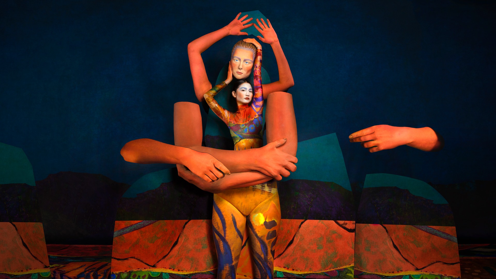 En kvinna håller en mask ovan sitt huvud, framför henne stora orangea rekvisita-armar.