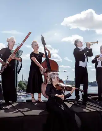 Flera musiker står på ett tak: de sminkade, bär peruker och bakom dem skiner solen.