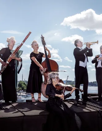 Several musicians stand on a roof: they are wearing make-up, wigs and the sun is shining behind them.
