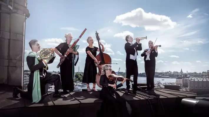 Flera musiker står på ett tak: de sminkade, bär peruker och bakom dem skiner solen.