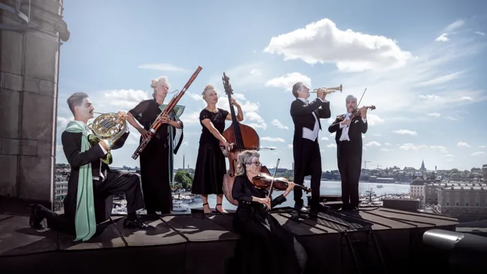 Flera musiker står på ett tak: de sminkade, bär peruker och bakom dem skiner solen.