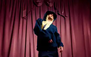 A man with long blond hair, wearing a dark hoodie, stands against a red curtain in the background.