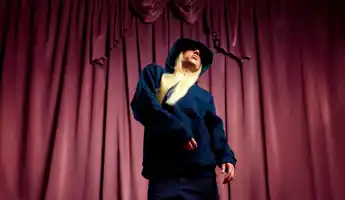 En man med långt blont hår, klädd i en mörk hoodie, står mot ett rött draperi i bakgrunden.