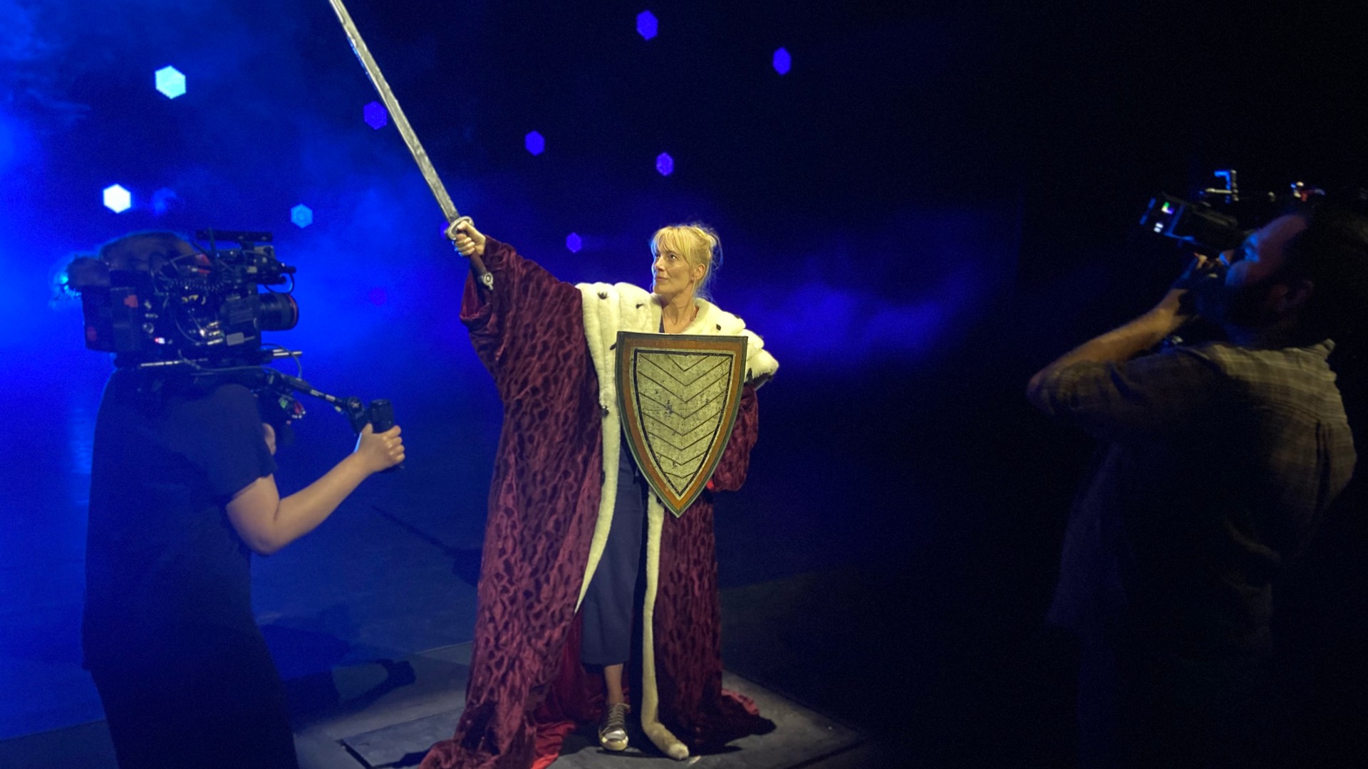På en scen med blått ljus står en kvinna med sköld och svärd, två män med kameror filmar.
