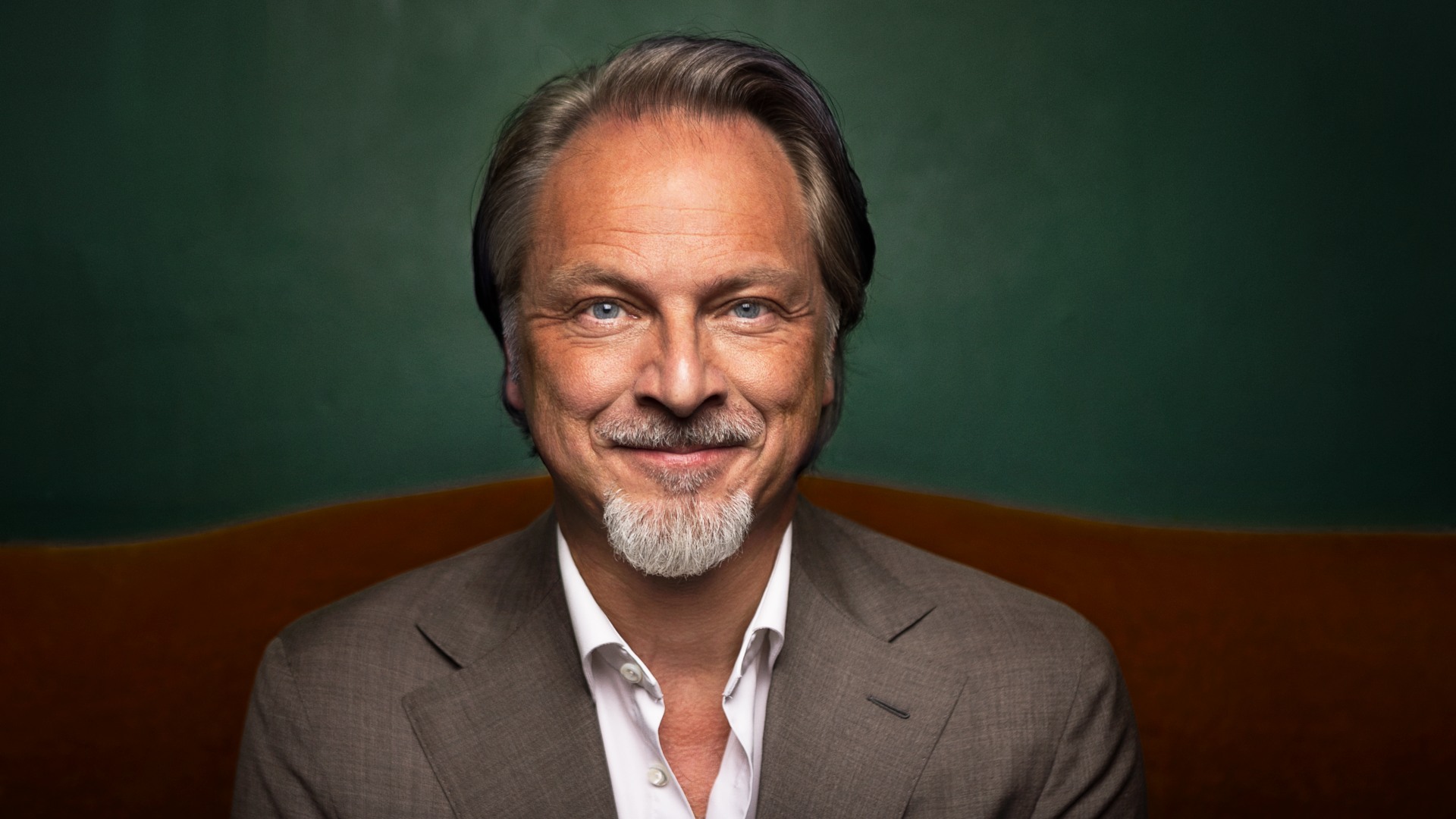 A smiling person in a grey suit and white shirt in front of a green wall.