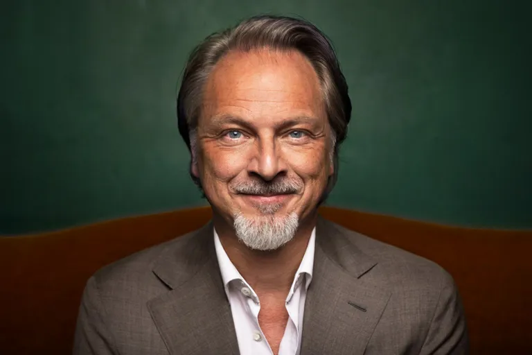 En leende person i grå kostym och vit skjorta framför en grön vägg.