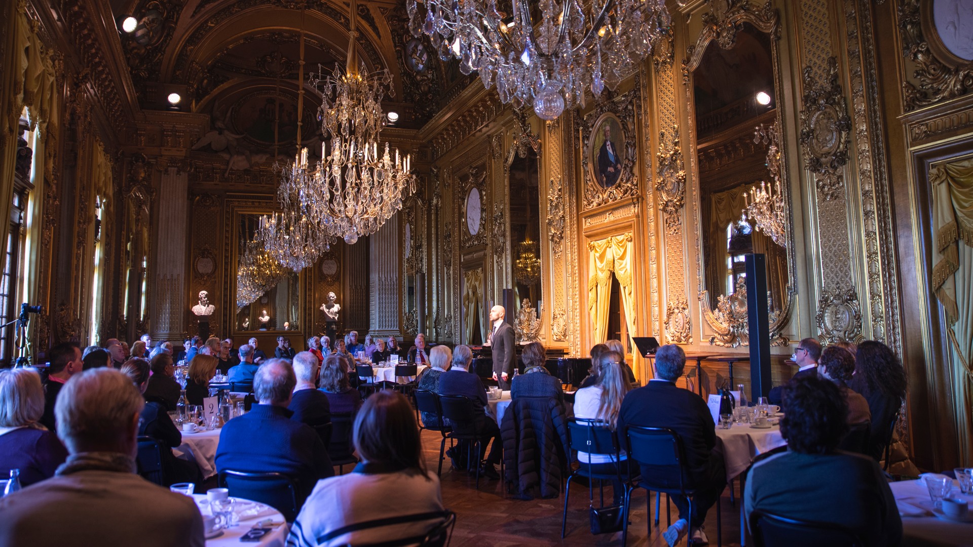 A gold-adorned room with large crystal chandeliers and several set tables and lots of people.
