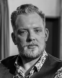 Porträttbild av en blond man med skägg och mustasch, han bär väst och en blommig skjorta.