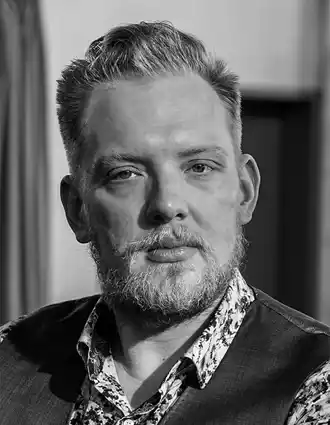 Porträttbild av en blond man med skägg och mustasch, han bär väst och en blommig skjorta.