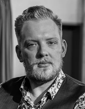 Porträttbild av en blond man med skägg och mustasch, han bär väst och en blommig skjorta.