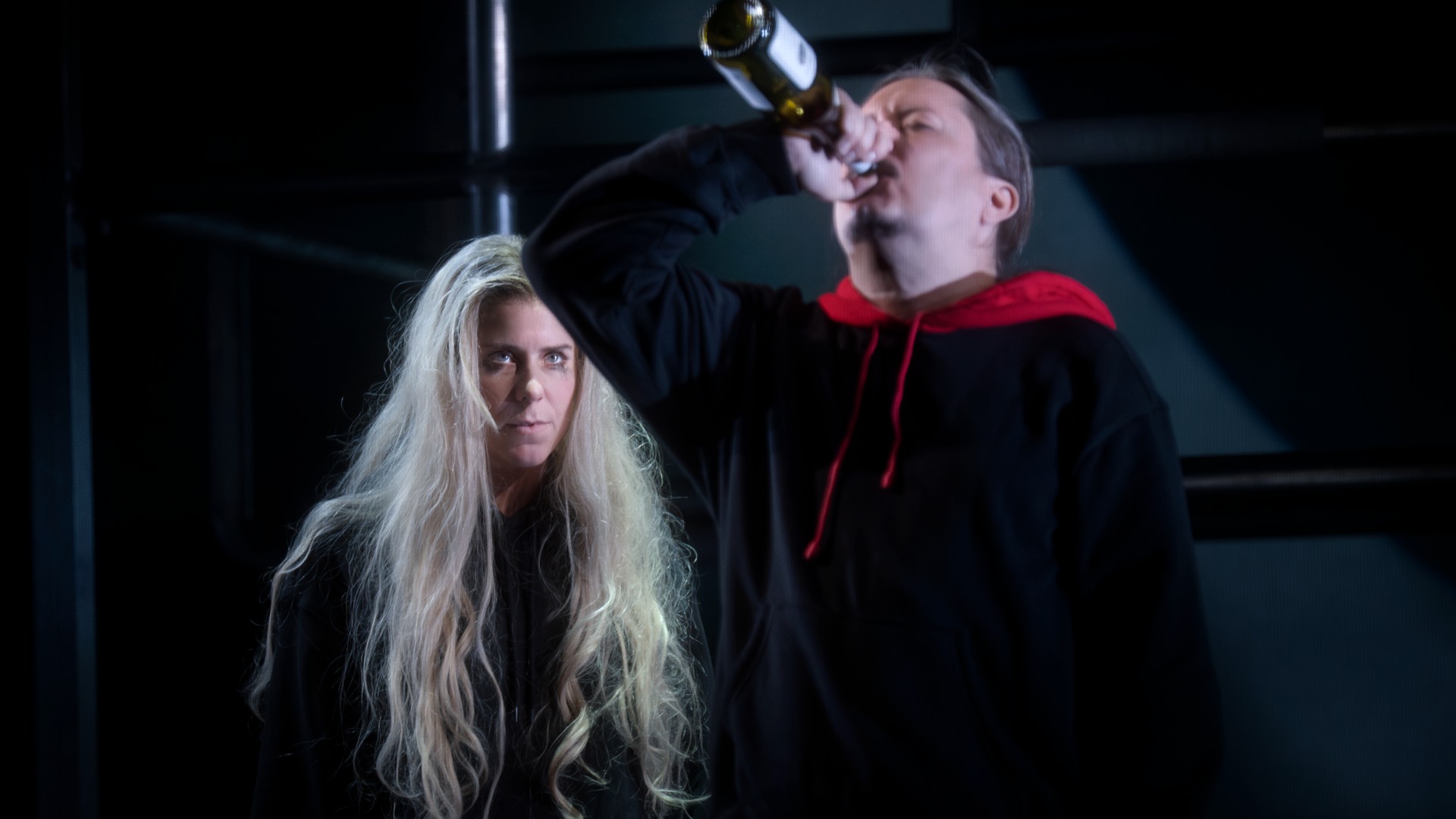 I förgrunden dricker en man vin direkt ur flaskan, i bakgrunden betraktas han av en långhårig kvinna.