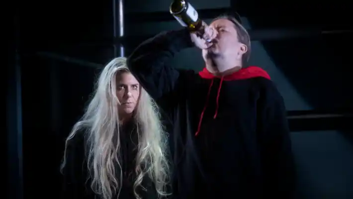 I förgrunden dricker en man vin direkt ur flaskan, i bakgrunden betraktas han av en långhårig kvinna.