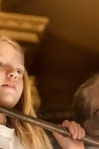 Two people with blurred faces stand behind a railing indoors, surrounded by warm tones.