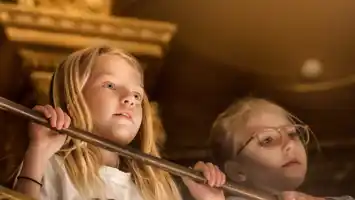 Två personer med suddade ansikten står bakom ett räcke inomhus, omgivna av varma toner.