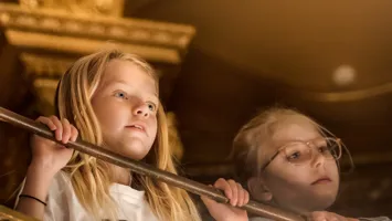 Två personer med suddade ansikten står bakom ett räcke inomhus, omgivna av varma toner.