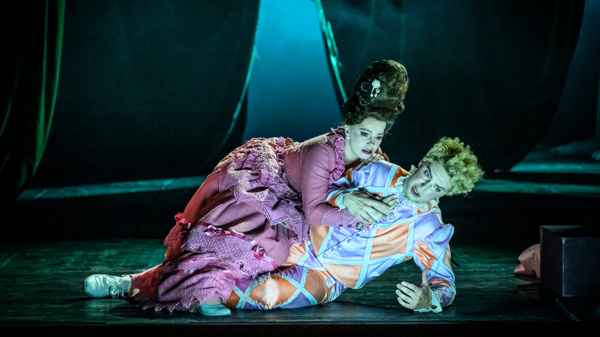 En kvinna i rosa klänning och en man i harlekinkostymer ligger på ett golv.