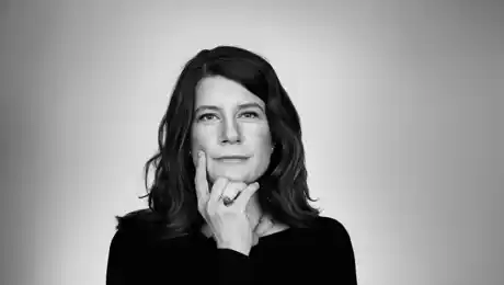 Ansiktsbild på kvinna med ring på höger hand och som gestikulerar vid munnen, mot enfärgad bakgrund.