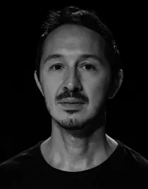 A close-up of a man with a beard and mustache in a dark shirt.