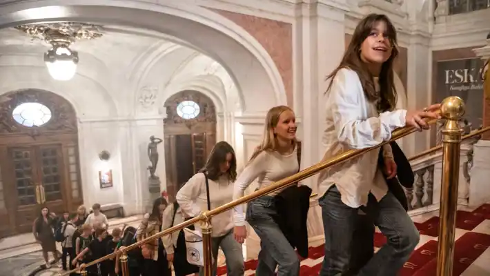 People walk on a staircase in an elegant building with arches and chandeliers.