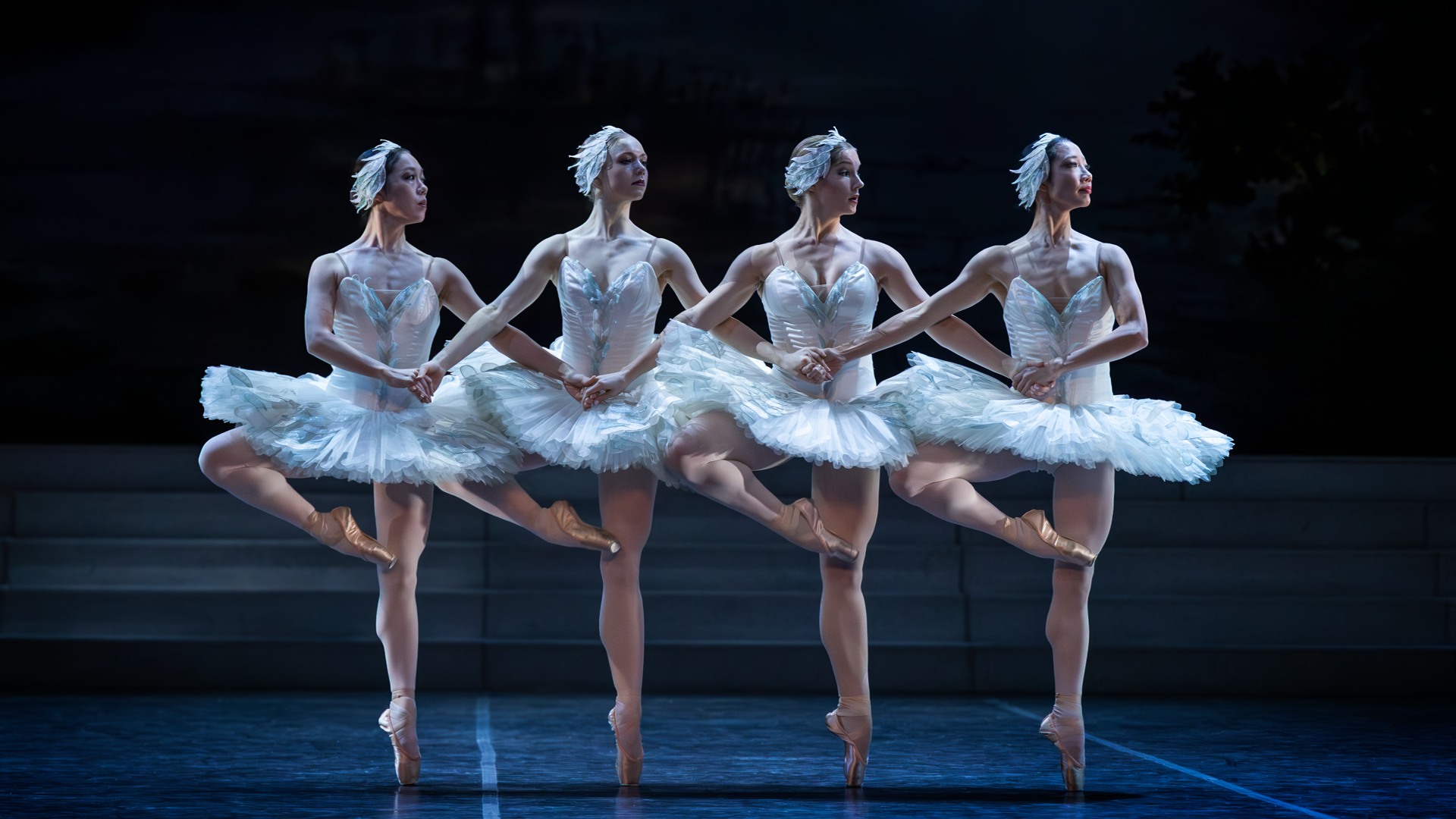Four dancers who look like swans dance in sync on a stage.
