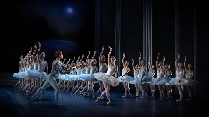 Many tulle-clad dancers who look like swans stand on a stage - a man with a bow and arrow approaches them.