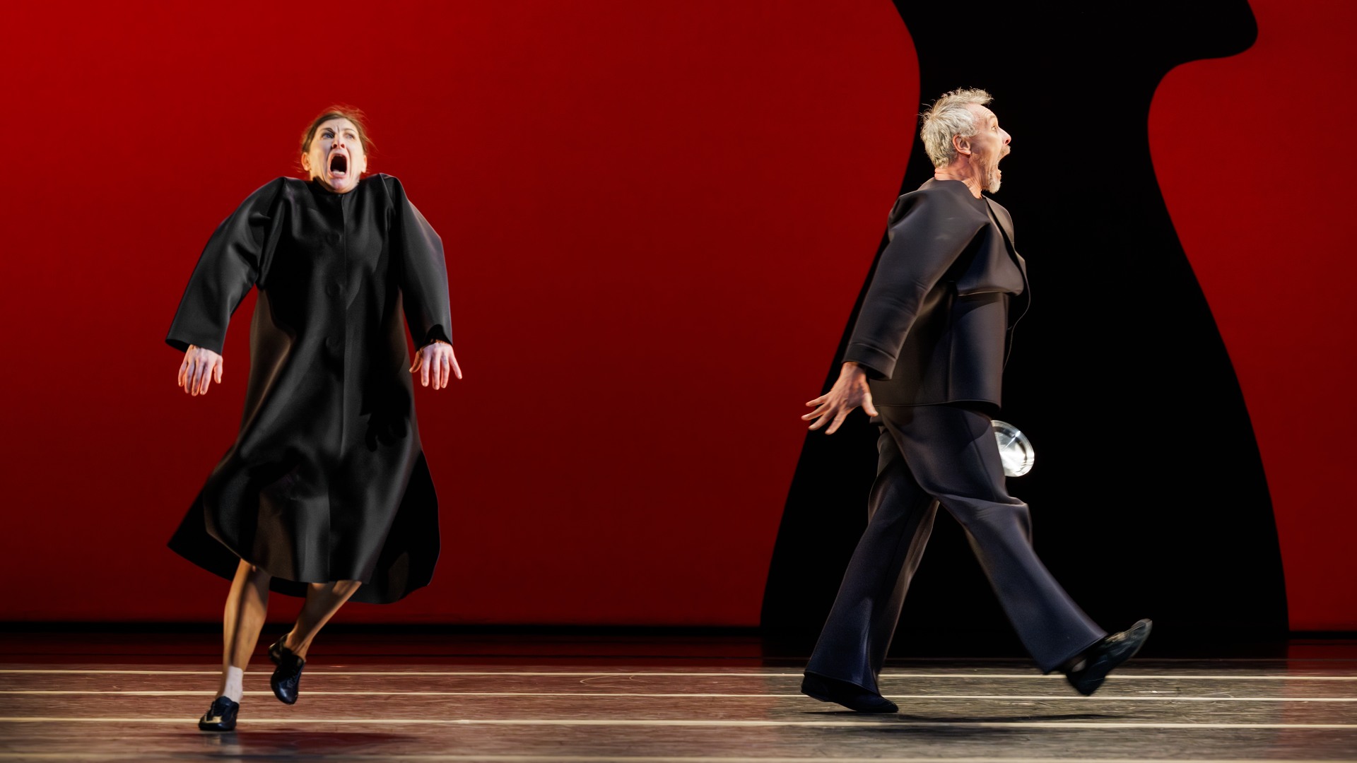 A woman and a man are moving stiffly in opposite directions against a red background.